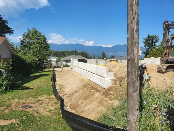 Construction site with a tiered retaining wall made of concrete blocks against a backdrop of mountains and blue sky.