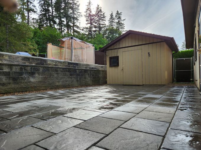 Patio with stone pavers, a wooden shed, and a low stone wall under a partly cloudy sky.