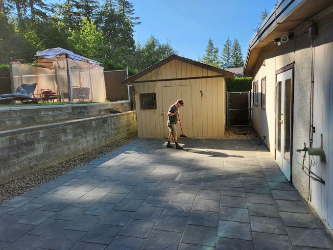 Person sweeping a paved backyard near a shed and house. Sunny day.