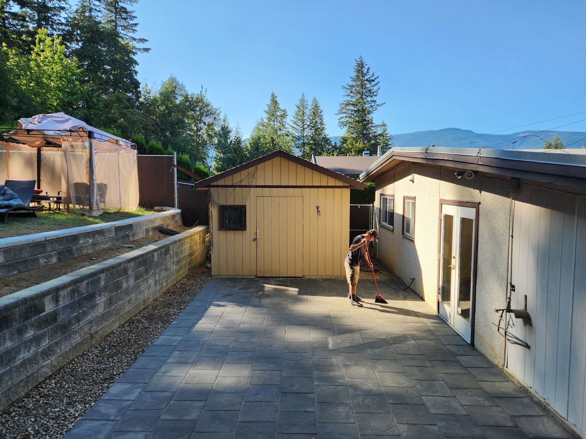 Backyard with a person sweeping, shed, building, and trees under a clear sky.