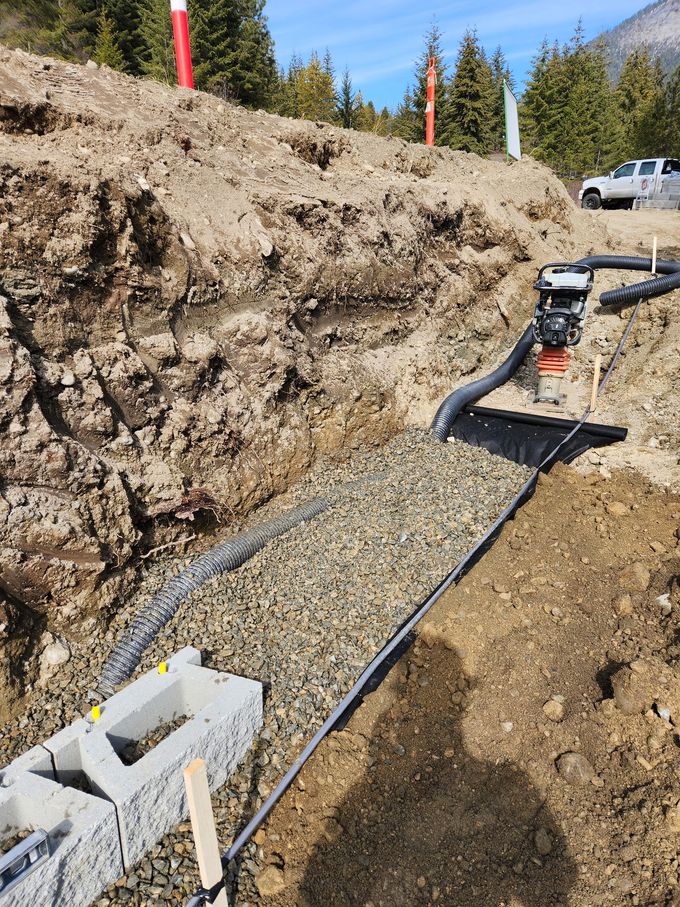 Construction site: trench with gravel, drain pipe, and a retaining wall, with a machine and hillside in the background.