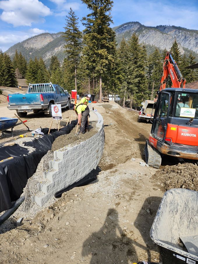 Construction site with workers building a retaining wall; blue truck, orange excavator, mountains in background.