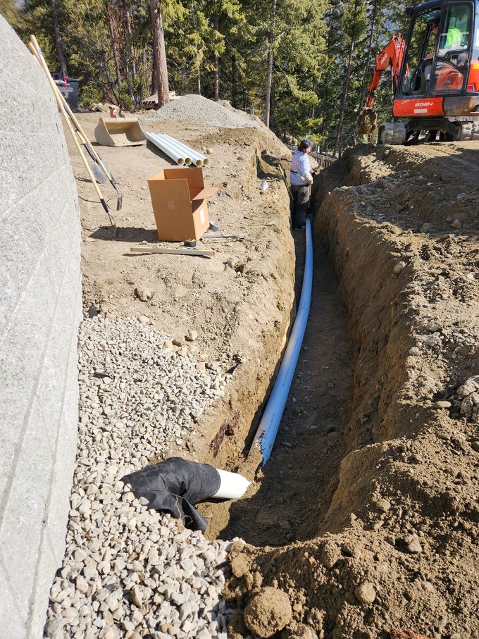 A trench with a white pipe and a person working on a construction site next to an excavator.