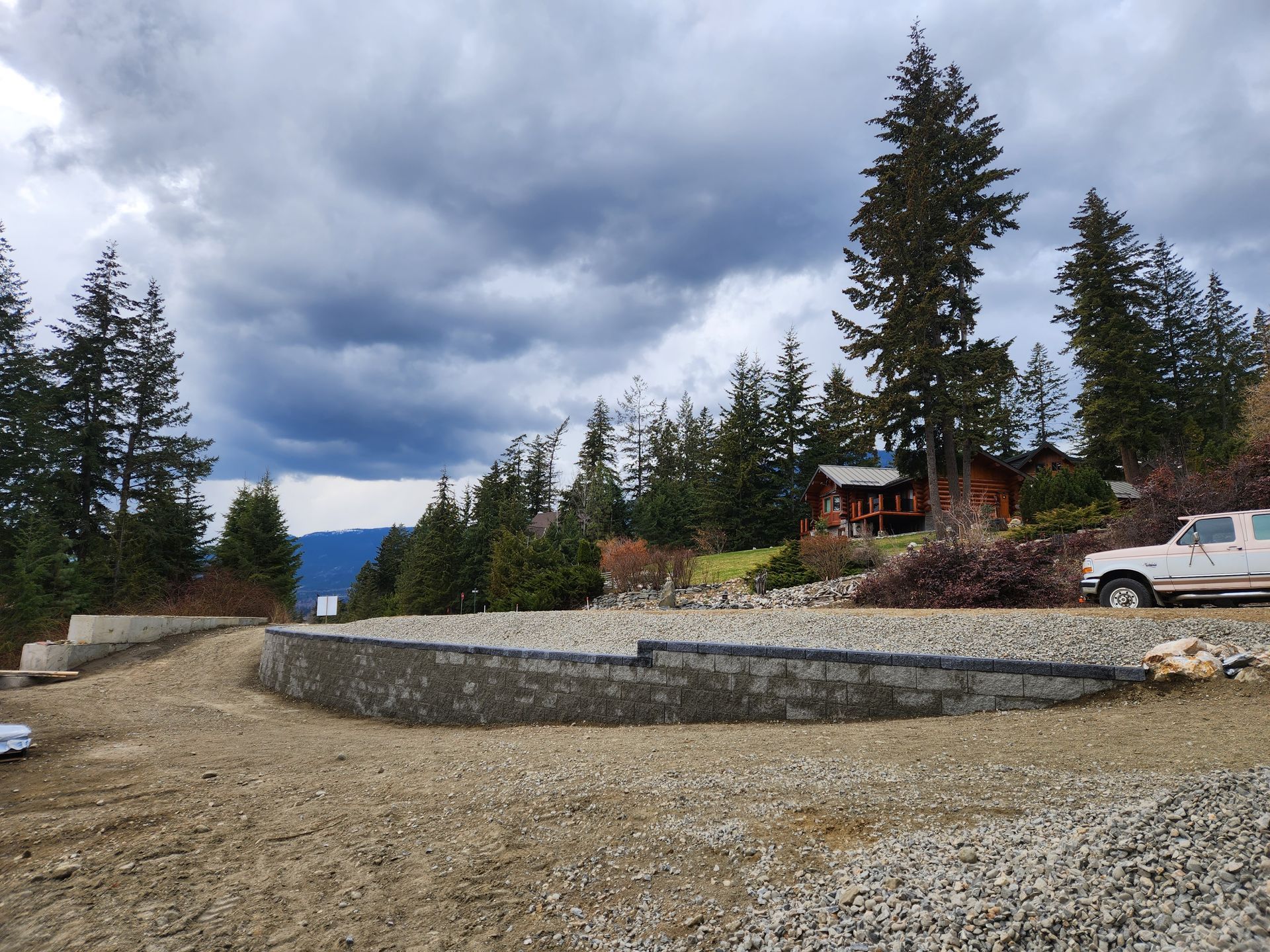 Gravel driveway with retaining wall, cabin and trees under cloudy sky. White pickup truck parked on right.