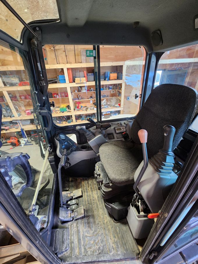 Interior of an excavator cab: seat, controls, windows; a workshop is seen through the back window.