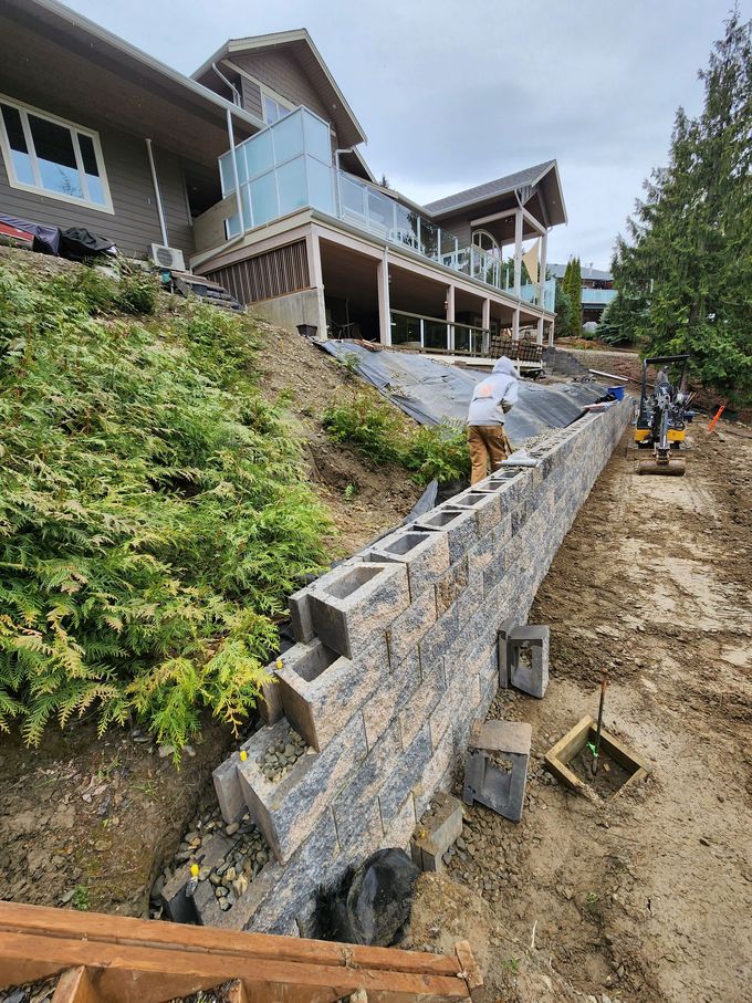 Retaining wall construction on a sloped yard near a house; a worker is placing concrete blocks.