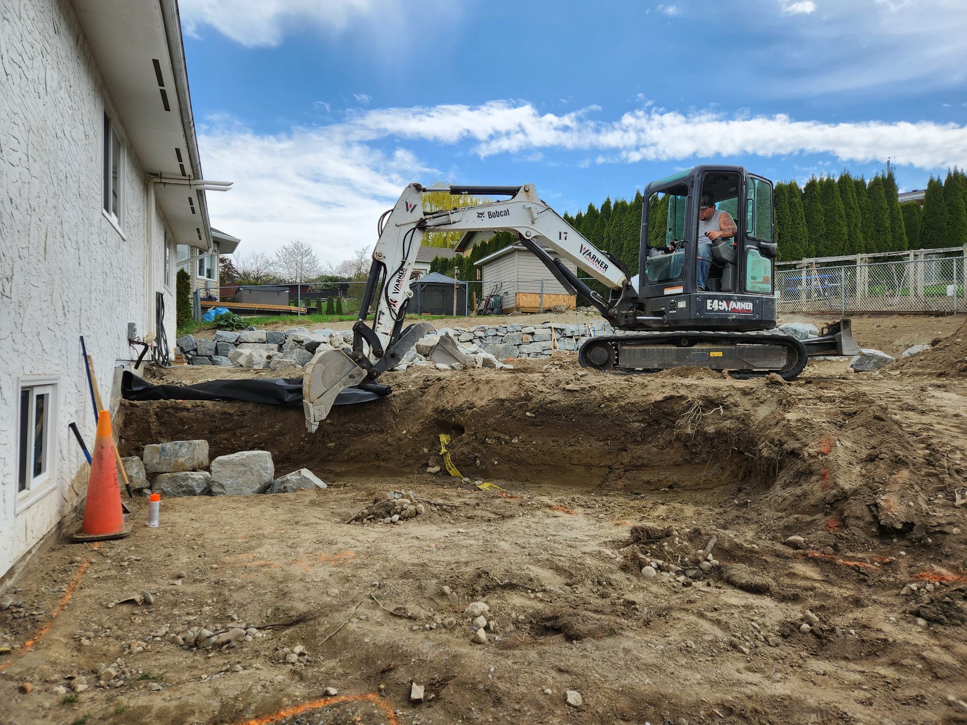 Mini excavator digs dirt near a house; an orange cone and rocks sit nearby under a blue sky.