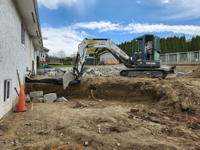 An excavator digging near a house. A worker is in the cab. Construction site with dirt, stone and a traffic cone.