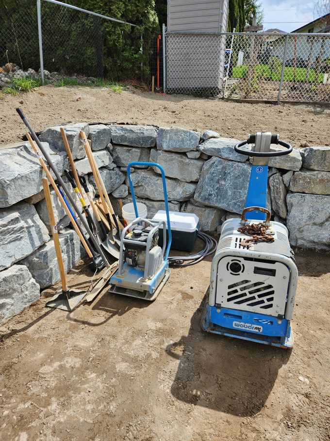 Two plate compactors and tools next to a stone retaining wall on a construction site.