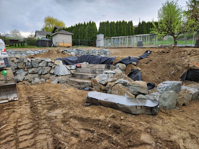 Construction site with stone retaining walls, concrete steps, and dirt. Cloudy day.