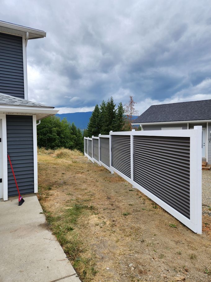White and gray fence with dark corrugated inserts beside a gray house, with a hilly, cloudy backdrop.