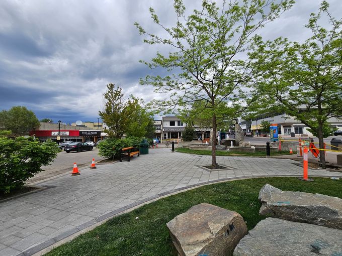 A paved plaza with trees and shops under a cloudy sky. Orange cones and rocks are in the foreground.