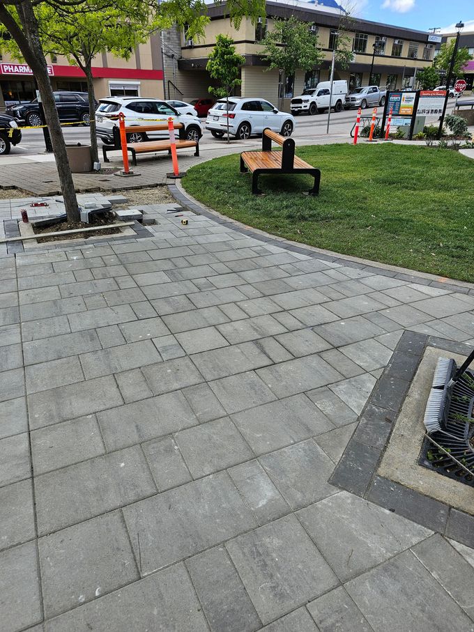 Sidewalk with gray pavers curves around a grassy area with a bench. Cars and buildings in the background.