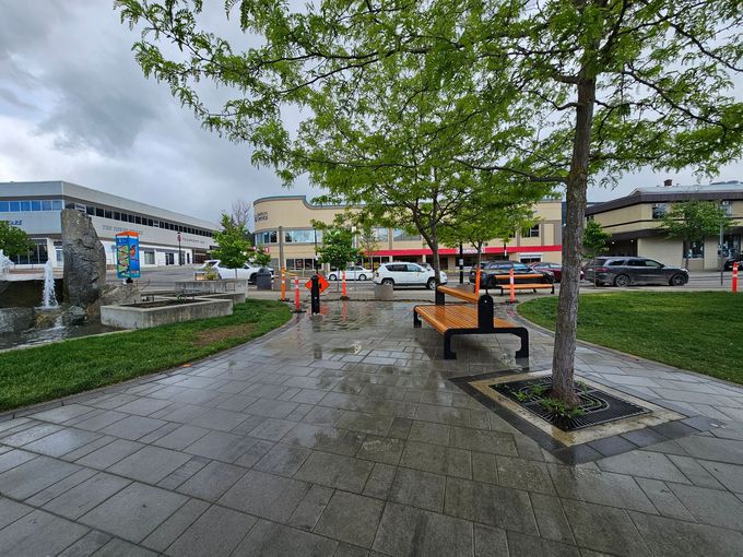 A paved plaza with a fountain, benches, and trees; buildings and cars are in the background on a cloudy day.