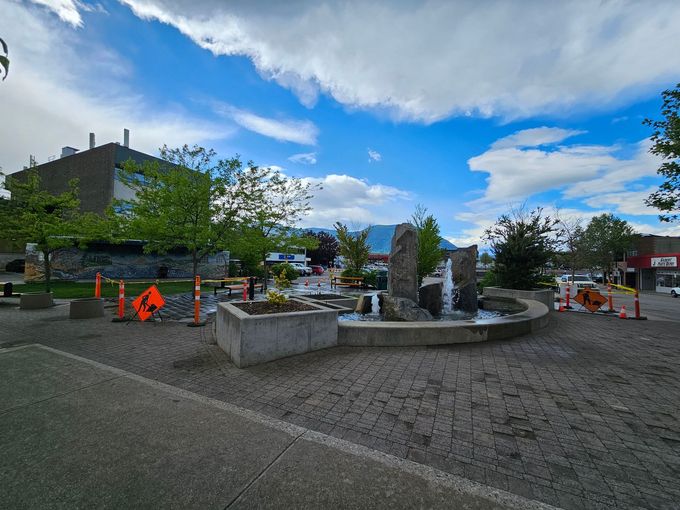 Fountain in a town square with three stone structures. Construction barriers surround the area. Blue sky above.