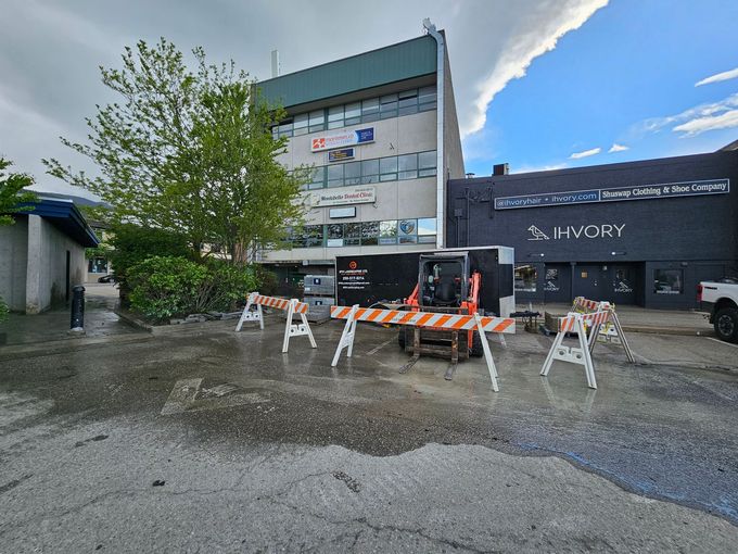 Construction site with barriers in front of a building.  Black machinery, gray asphalt, cloudy sky.