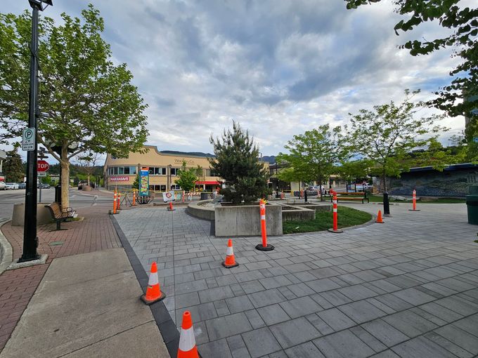 Paved town square with orange cones around a fountain. Trees, buildings, and cloudy sky visible.