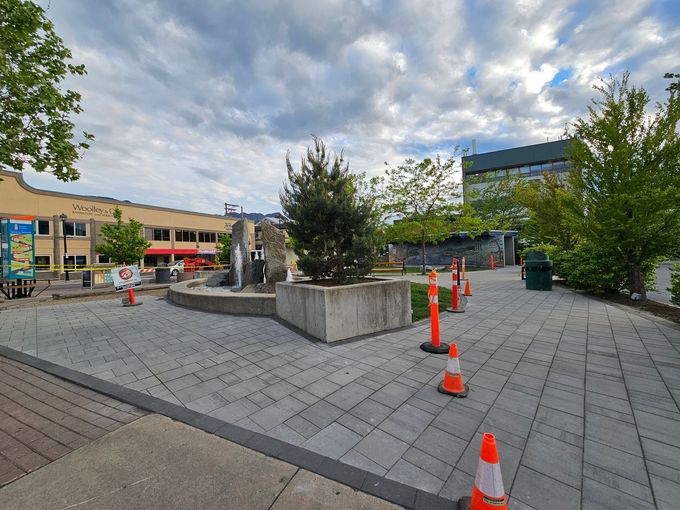 Plaza with a fountain, trees, orange cones, and buildings under a cloudy sky.