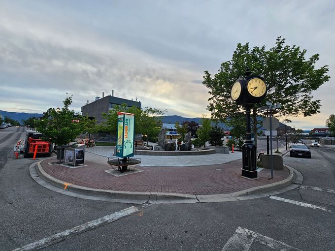 Clock tower in a roundabout with trees, signs, and buildings under a cloudy sky.