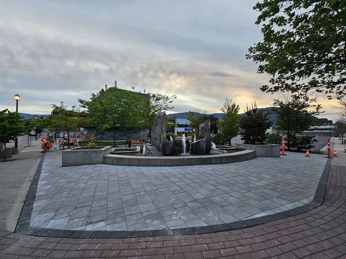 Fountain with stone sculptures and water spouts, set in a paved plaza with trees. Cloudy sky in background.
