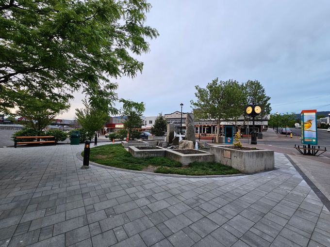 Fountain and plaza in a town square, trees, benches, shops, and a clock tower. Overcast sky.