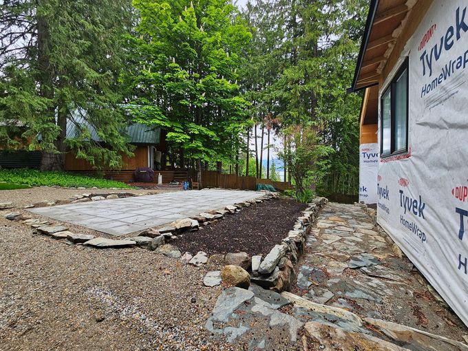 A partially built house with Tyvek wrap next to a garden bed with gravel and trees in the background.