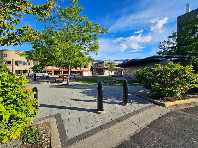 A park square with trees, gray brick ground, buildings in the background, and a blue sky with white clouds.