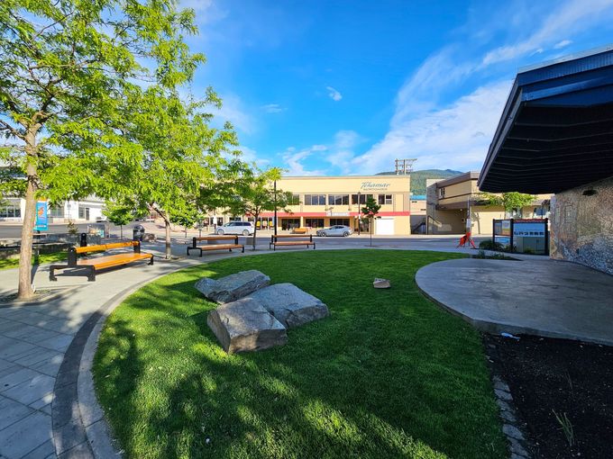 Park scene with green lawn, benches, and buildings under a blue sky.