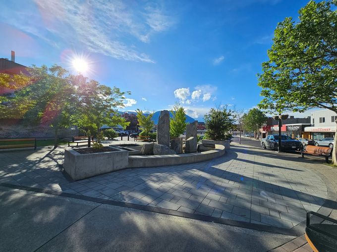 Sunny town square with trees, stone sculpture, and street. Blue sky with some clouds.