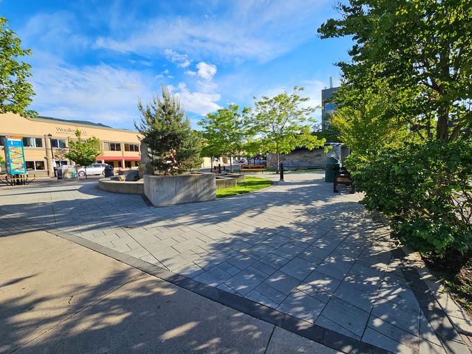 A sunny plaza with gray pavers, greenery, and buildings under a blue sky.