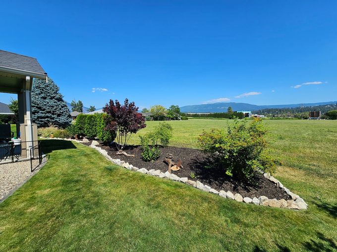 A sunny backyard with a garden bed, lush grass, and a view of distant hills.