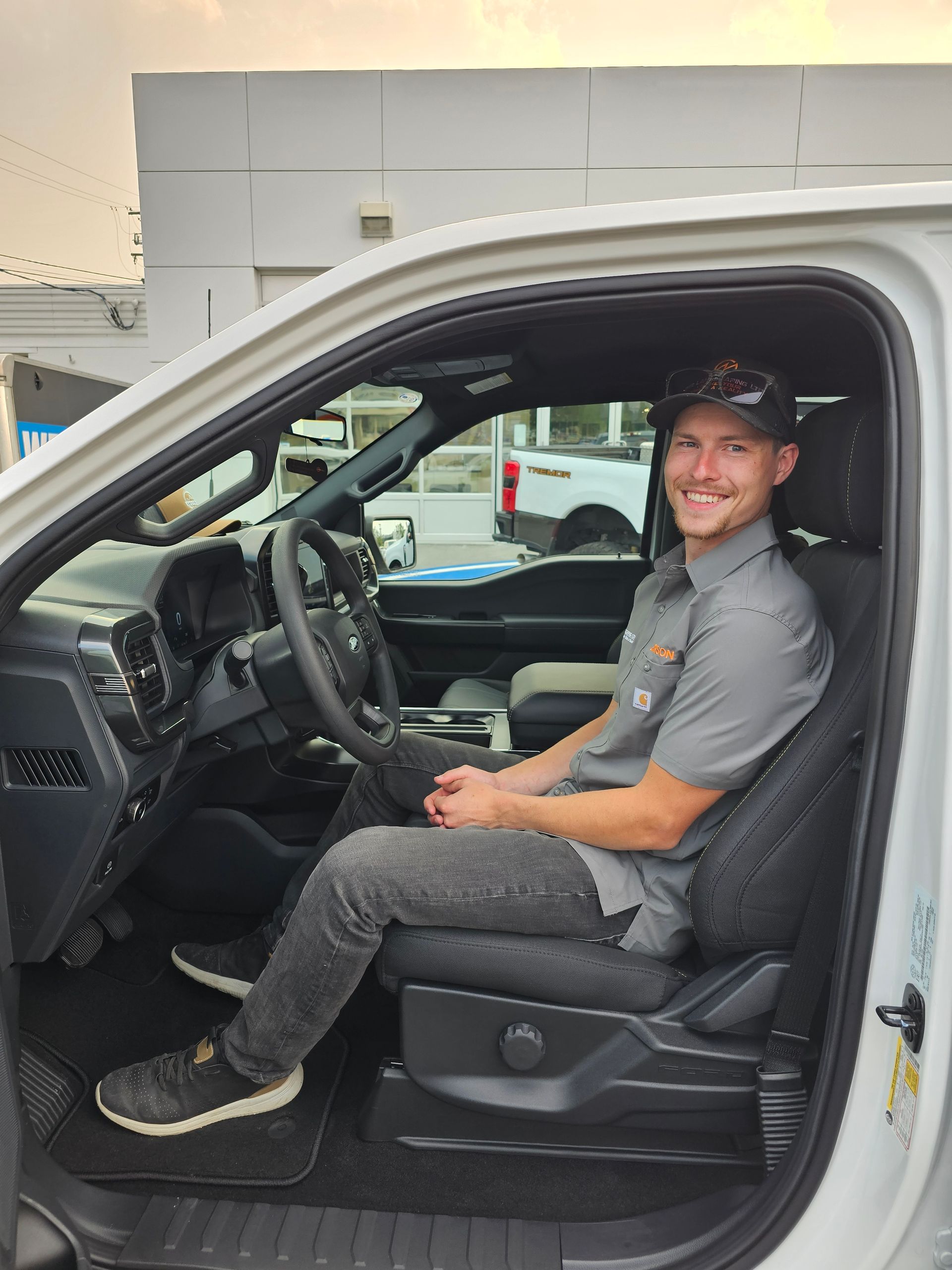 Man sitting in the driver's seat of a white pickup truck, smiling. He wears a gray uniform and a baseball cap.