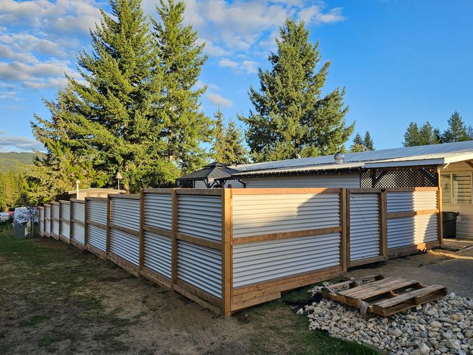 A wooden fence with corrugated metal panels encloses a yard. Trees and a house are in the background.