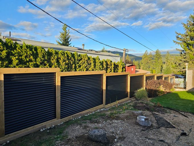 A wooden fence with black wave-like panels in a backyard with a view of trees and sky.