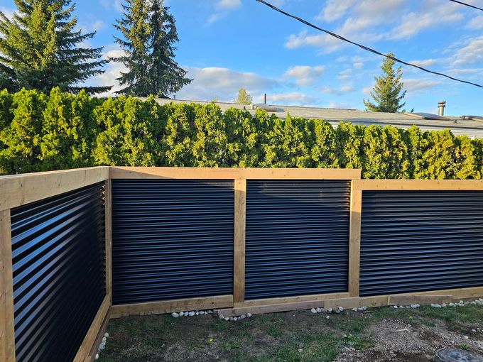 A black corrugated metal fence with a wooden frame in front of a green hedge under a blue sky.