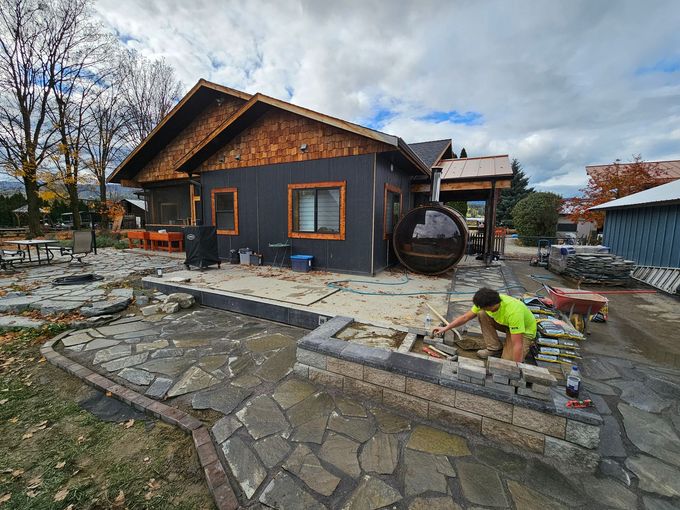 Man laying stone in front of a dark-sided building with a wooden sauna. Overcast day.