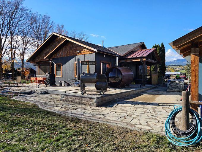 Cabin with a wooden barrel sauna, sitting on a stone patio with blue sky and mountains in the background.