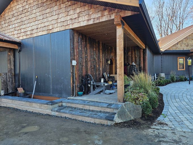 Exterior of a home with wood and dark siding, covered porch, stone steps, and circular brick driveway.