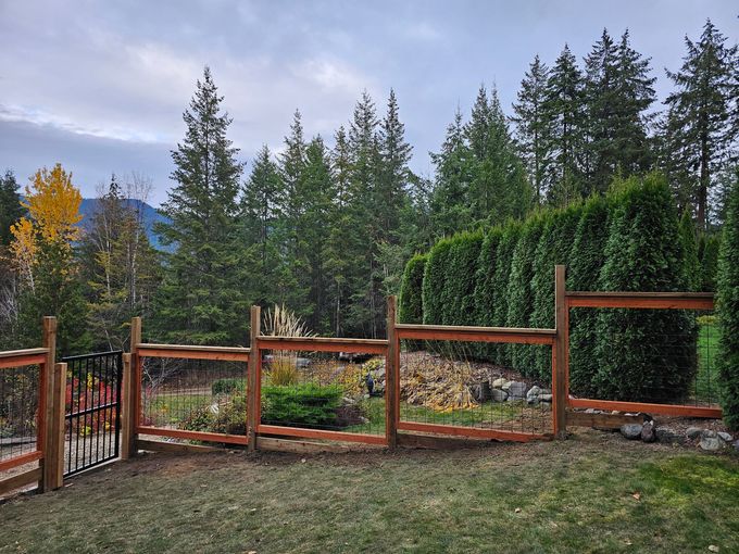 A wooden fence with a gate encloses a yard, lush with greenery, backed by trees and a mountain.