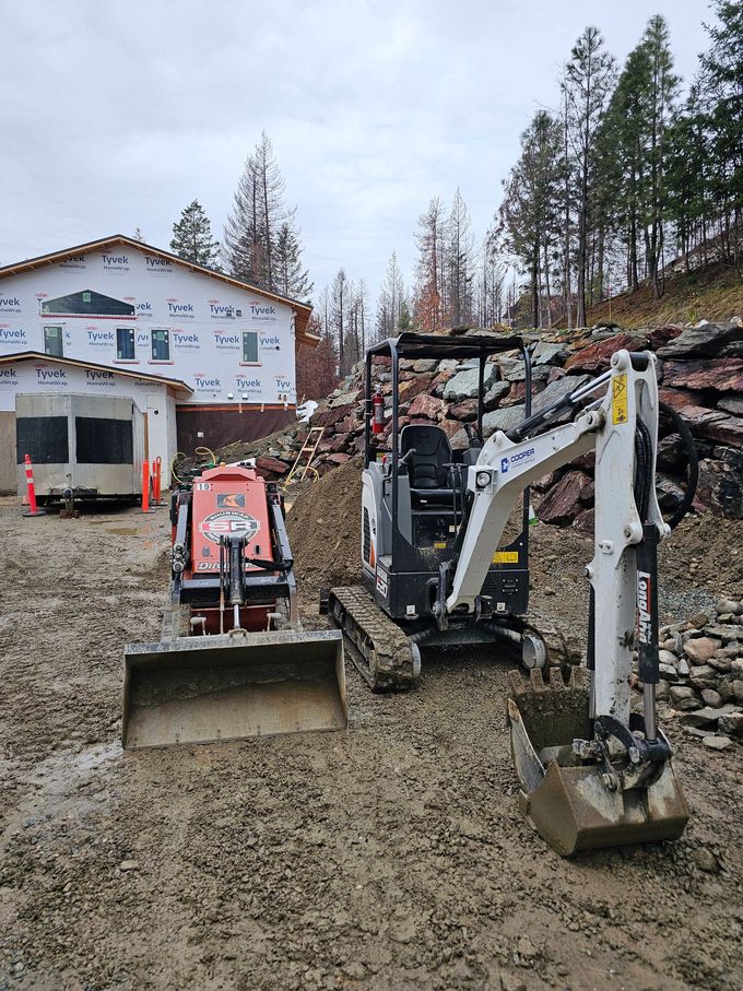 Two small excavators on a muddy construction site with a partially built house and forested hillside.