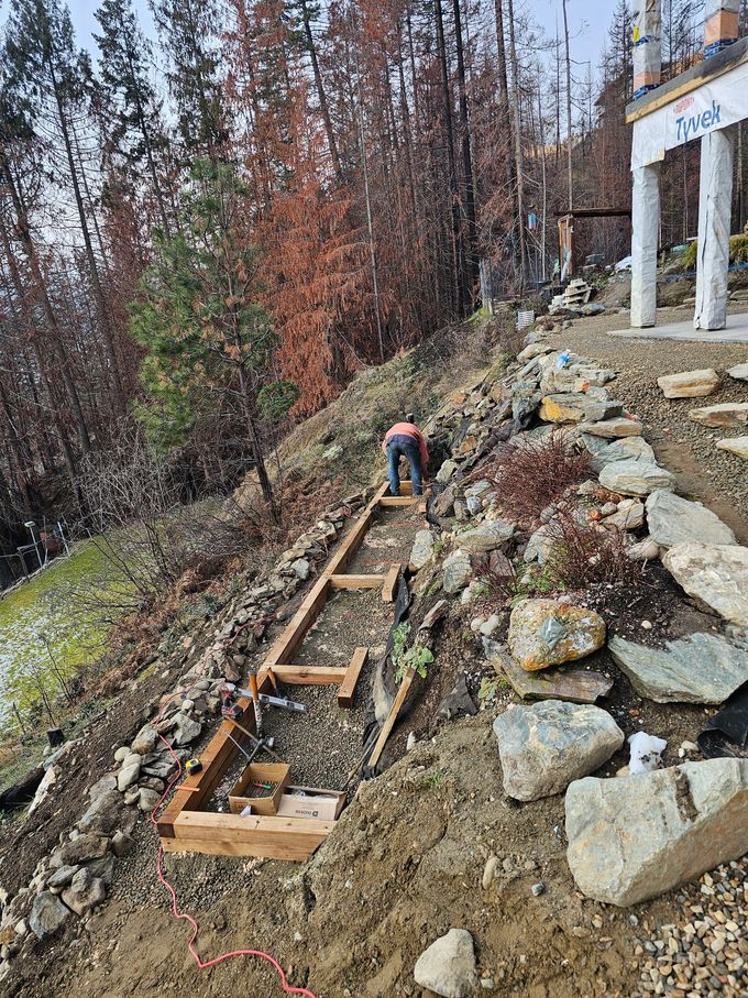 Construction worker building wooden steps on a steep, rocky hillside. Earth tones dominate, with a white building visible.