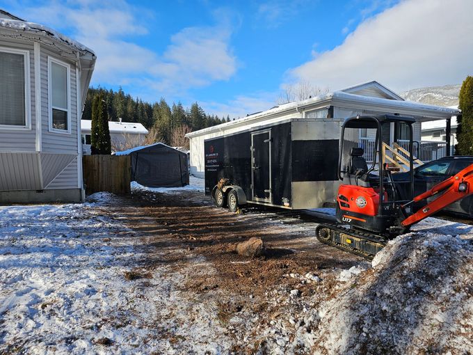 A construction scene with a trailer, excavator, snow, and houses on a sunny day.