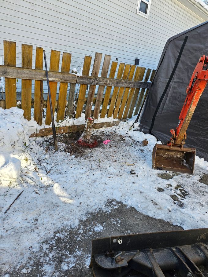 Mini excavator digging in snow next to a weathered wooden fence.