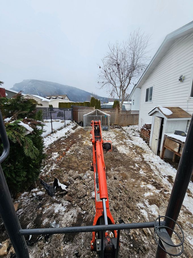 Mini excavator in a snowy backyard, digging near a fence and house.