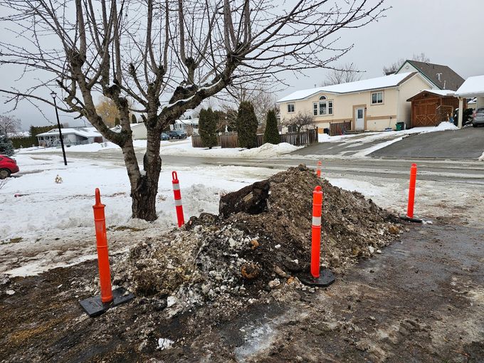Pile of debris on a snowy street, surrounded by orange traffic cones, near a tree and houses.