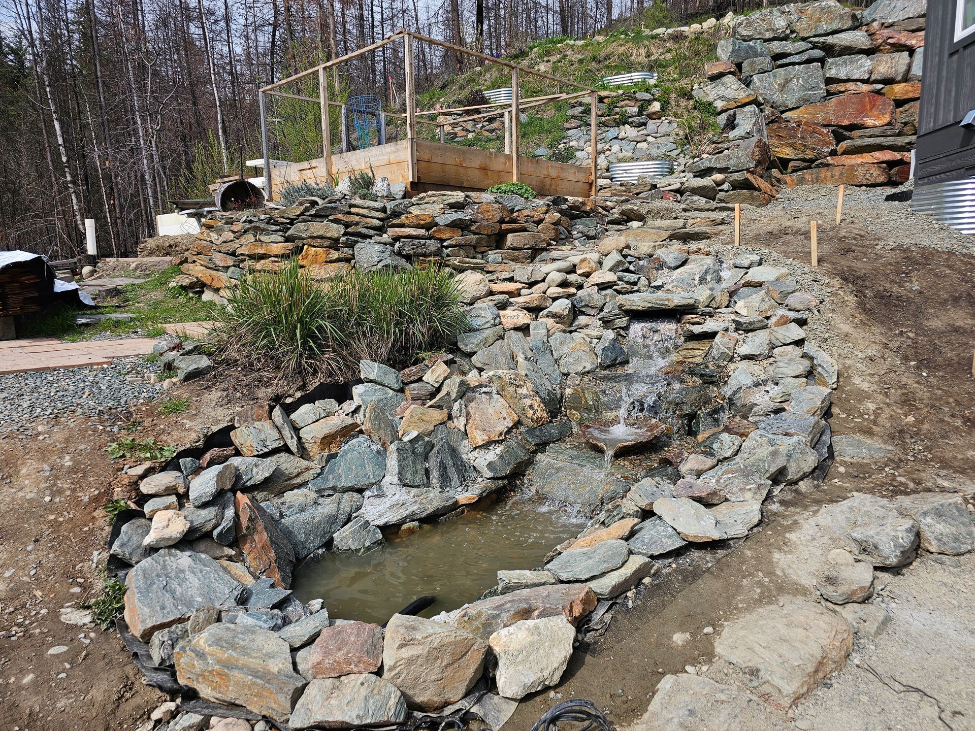 Rock waterfall in a garden, flowing into a small pond, with wooden structure in the background.