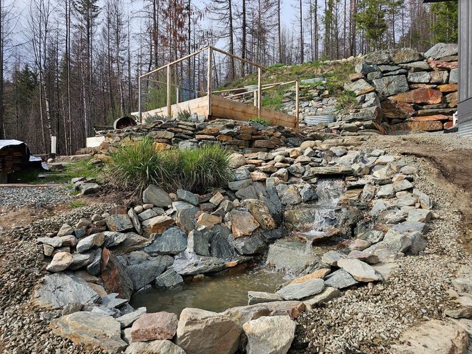 A water feature cascades over rocks in a landscaped yard, with a wooden structure visible behind.