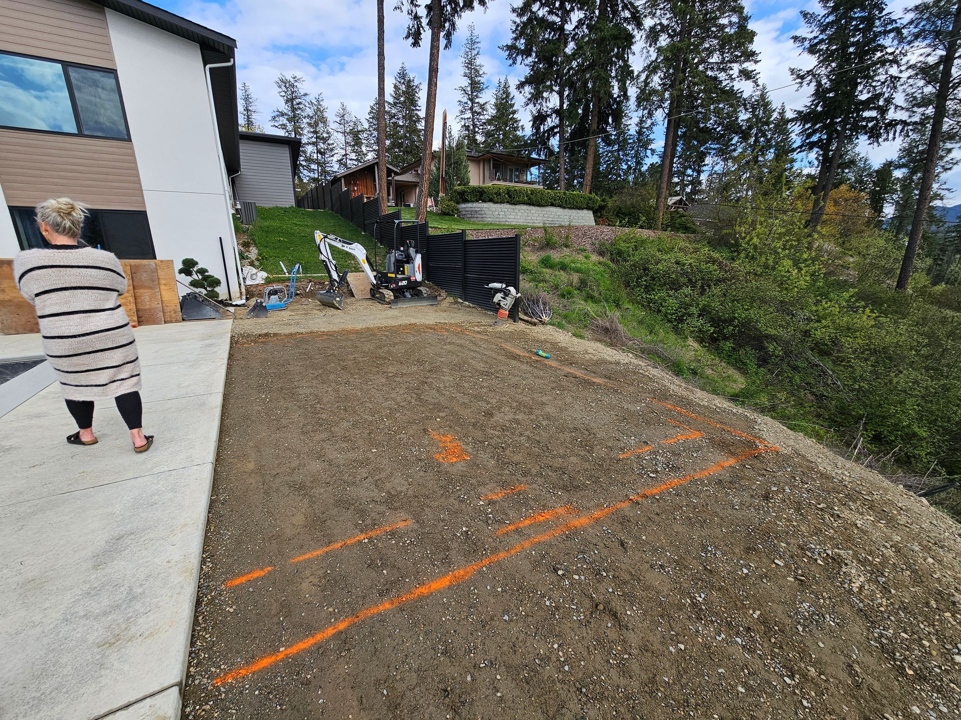 Woman standing near a dirt lot with orange lines, construction equipment, and a hillside with trees.