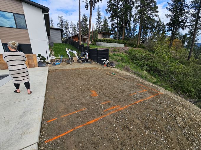 Woman standing near a dirt lot with orange lines, construction equipment, and a hillside with trees.