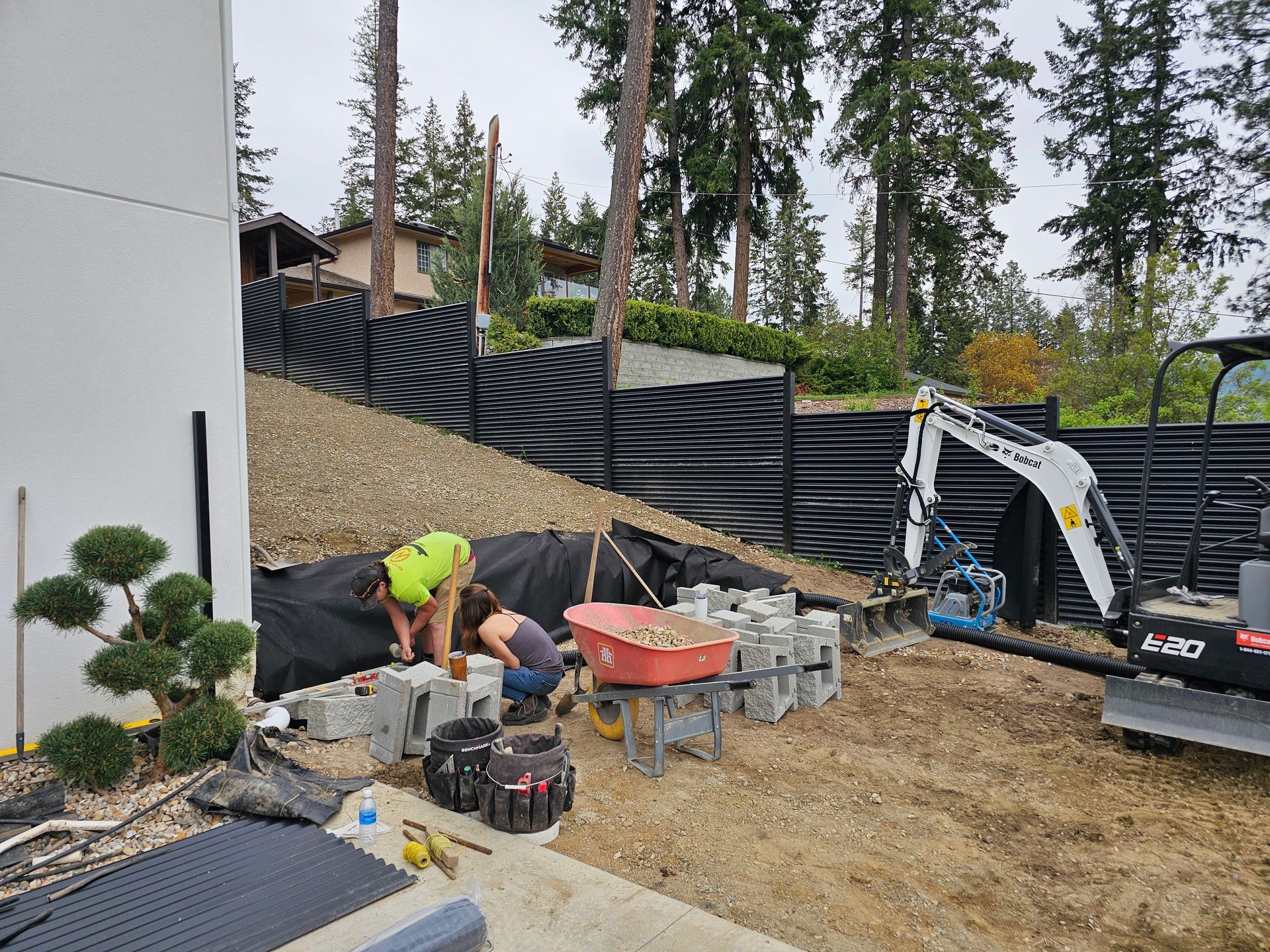 Two people constructing a retaining wall with blocks, hillside backdrop, small excavator.